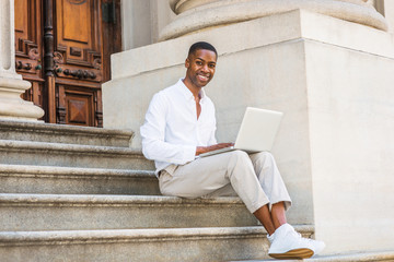 Way to Success. African American College Student studying in New York, wearing white shirt, light gray pants, sneakers, sitting on stairs outside office on campus, smiling, working on laptop computer