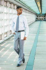 Fototapeta na wymiar Young African American Businessman working in New York, wearing white T shirt, light gray pants, black leather shoes, tie, holding laptop computer, standing on walkway in office building, looking away