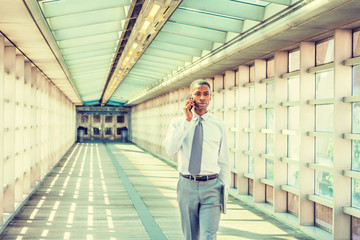 Young African American Businessman working in New York, wearing white shirt, necktie, holding laptop computer, walking on walkway with glass walls, ceiling, wooden floor, talking on cell phone..