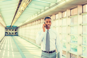 Fototapeta premium Young African American Businessman working in New York, wearing white shirt, necktie, holding laptop computer, walking on walkway with glass walls, ceiling, wooden floor, talking on cell phone..
