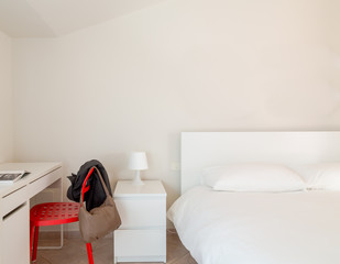Bedroom. White interior and simple decoration. White bed sheets, white  nightstand and white lamp. A jacket and a bag on a intese red chair