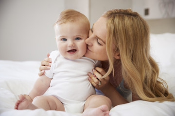 Mother Lying On Bed At Home Gives Baby Son Kiss
