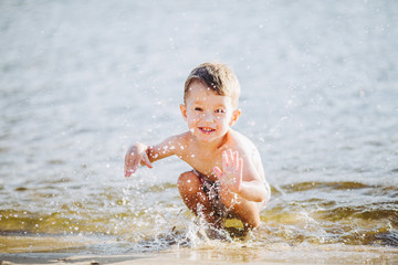 boy child playing makes splashes, beats hands on the water in the river at sunset
