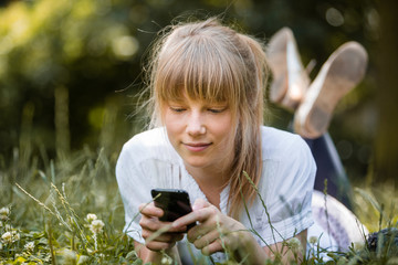 Young woman with fringe lying down on her stomach on grass with mobile phone