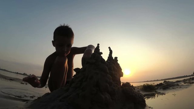 Silhouette of a Child on the Beach Building a Sand Castle at Sunset. Happy little boy sitting on the river bank and plays with sand. Summer vacation.
