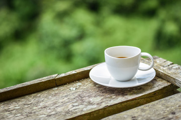 White coffee cup placed on wooden bar, view of bright morning sunlight and green garden background. 