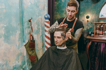 Close up shot of man getting trendy haircut at barber shop. Male hairstylist in tattoos serving client.