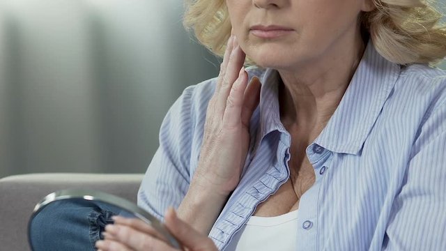 Elderly woman holding mirror, looking at neck and touching, flabby skin, lifting