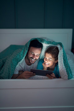 Beautiful smiling couple lying on the bed under blanket and watching internet content on a tablet.