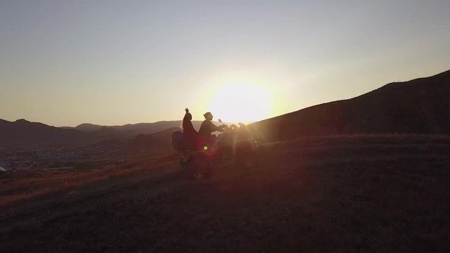 Aerial shooting from flying drone of a young couple of tourists is enjoying beautiful sea landscape and hills on sunset in slowmotion, while is riding on a ATV transport or quad bike in 4K