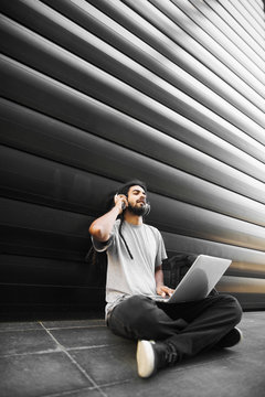 Young bearded hipster man with headphones enjoying in music while sitting on the street with a laptop.
