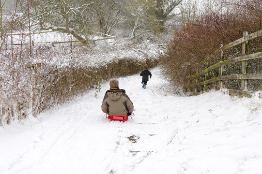Child runs down snow slope with father chasing behind on sled