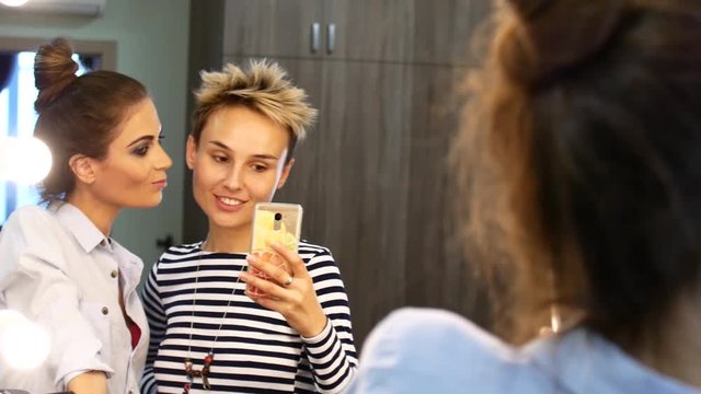 Two friends make a selfie near the mirror. Girls fool around laughing and taking pictures.