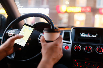 Driver with a smartphone in hand and paper cup of hot coffee in the driver's seat. The concept of inattention at the wheel, rest, coffee break to cheer. Man paves way in gps. On screen map of area.