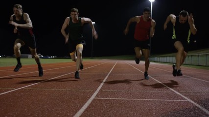  Competitive male athletes running at race track in the dark. Slow motion.