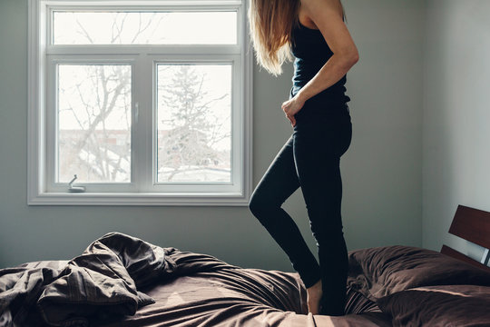 Blonde woman in a bedroom standing on a bed with rumpled sheets.