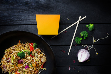 Udon stir fry noodles with chicken and vegetables in wok pan on black wooden background. With a box for noodles