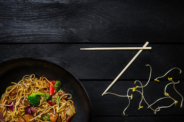Udon stir fry noodles with chicken and vegetables in wok pan on black wooden background. With chopsticks