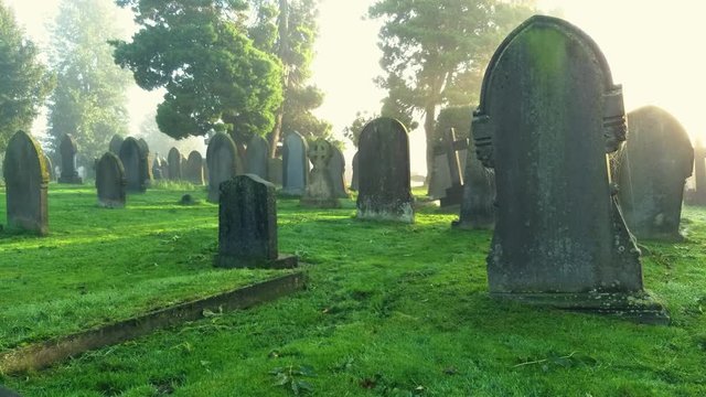 Misty morning sun on old graveyard brigs hope with new rays of light. Weathered gravestones in fog at a cemetery. Static camera , moisture particles moving.