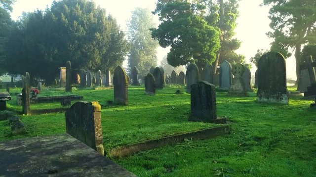 Misty morning sun on old graveyard brigs hope with new rays of light. Weathered gravestones in fog at a cemetery. Static camera , moisture particles moving.