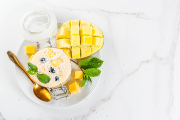 Dietary drink, breakfast. Tropical mango smoothie with fresh pieces of mango, blueberries, coconut and mint leaves. In a glass jar, on a white marble table. Copy space top view
