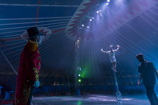 Circus performer on unicycle on stage with ring master and stage hand