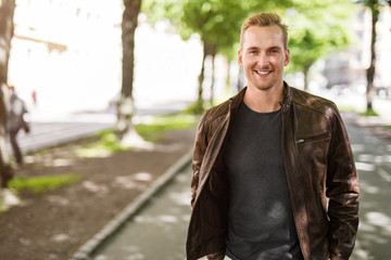 Trendy blonde man standing outdoors in a city, with trees around him wearing a brown leather jacket smiling towards camera.