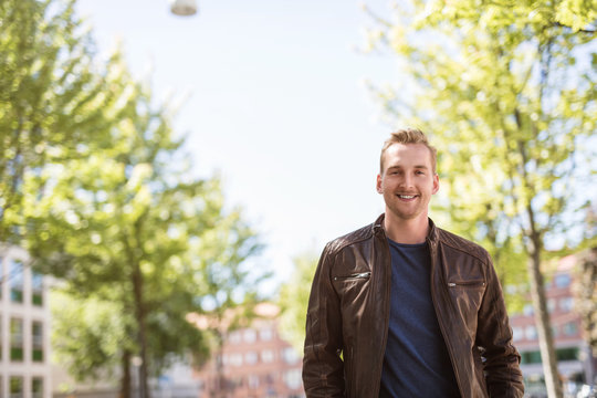 Trendy blonde man standing outdoors in a city, with trees around him wearing a brown leather jacket smiling towards camera.