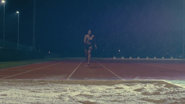  Female long jumper training at athletics track in the dark & rain. Slowmo
