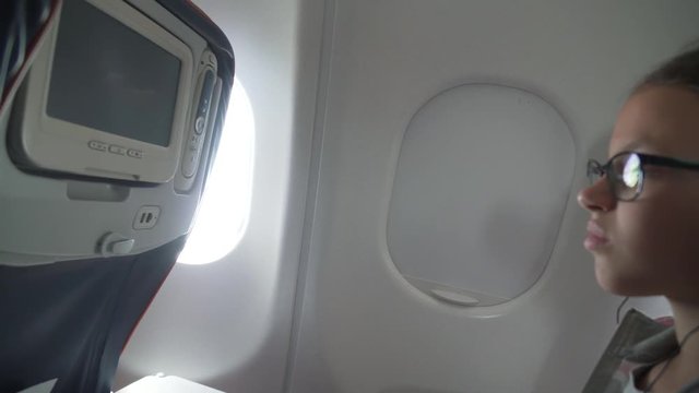 Young girl with glasses and headphones watches video on the monitor built into armchair in the cabin of the airplane stock footage video