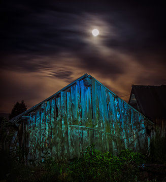 A spooky old wooden barn at night illuminated with full moon and stars glowing through overcast skies in background.