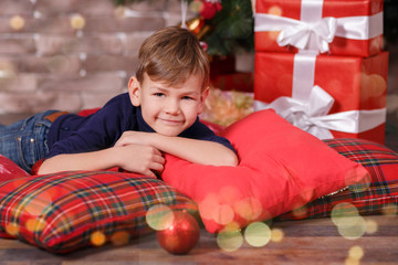 Fototapeta na wymiar Handsome cute boy celebrating New Year Christmas alone close to xmas tree on red pillow posing in studio decoration wearing jeans and blue shirt with jazzbow tie