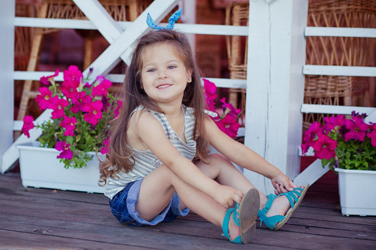 Stylish beautifull cute baby girl with brunette hair posing on wooden garden full of flowers wearing tiny jeans shirts and airy skivy underwaist and blue sandals