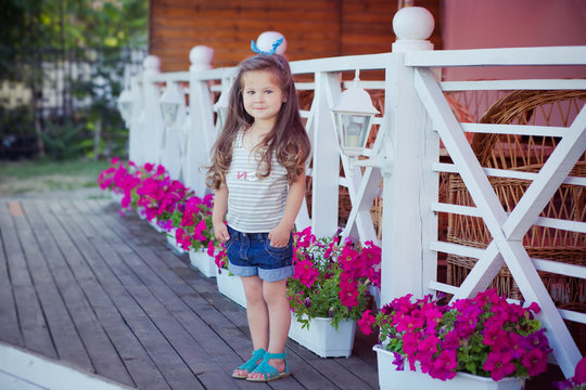 Stylish beautifull cute baby girl with brunette hair posing on wooden garden full of flowers wearing tiny jeans shirts and airy skivy underwaist and blue sandals