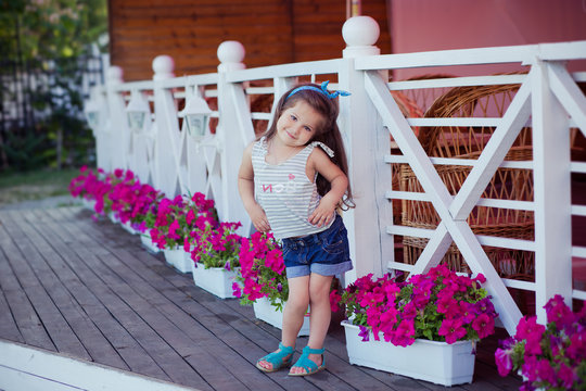 Stylish beautifull cute baby girl with brunette hair posing on wooden garden full of flowers wearing tiny jeans shirts and airy skivy underwaist and blue sandals