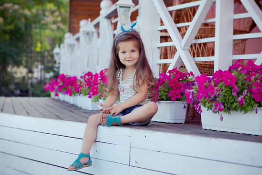 Stylish beautifull cute baby girl with brunette hair posing on wooden garden full of flowers wearing tiny jeans shirts and airy skivy underwaist and blue sandals