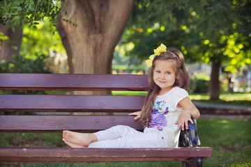 cute musical baby child girl with brunette hairs and stylish wear enjoying life sitting on wooden chair bench in summer awesome park alone