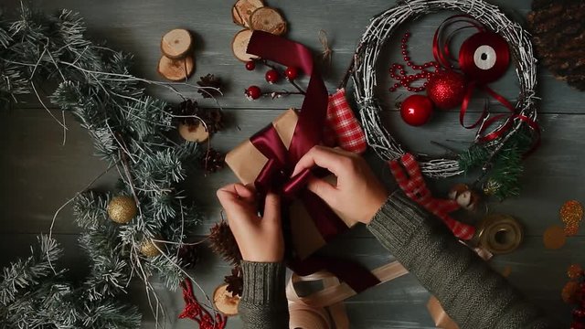 A top-down plan. Fully visible the table with the decorations. Female hands put and finalize Christmas gift wrapped in craftool paper on a wooden table. Bandaging tape and tied in a bow.