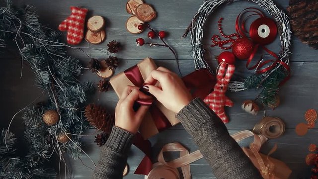 A top-down plan. Fully visible the table with the decorations. Female hands put and finalize Christmas gift wrapped in craftool paper on a wooden table. Bandaging tape and tied in a bow.