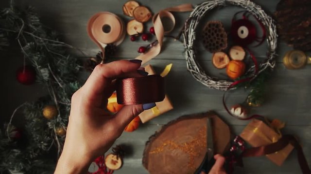 A top-down plan. Fully visible the table with the decorations. Female hands put and finalize Christmas gift wrapped in craftool paper on a wooden table. Bandaging tape and tied in a bow.
