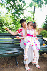 The young mother sitting on bench outdoor and keeping on the laps her beautiful daughter who is...