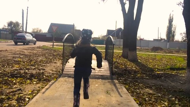 Young beautiful girl run away on park footbridge, slow motion. Small long bridge at picturesque autumn park, cross channel, high trees on background, flying birds.