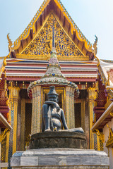 Fototapeta premium The famous Buddhist temple Wat Phra Kaew, located in the The Grand Palace in the historic centre Bangkok. In the foreground, the statue of the Seated Hermit, behind, the temple of the Emerald Buddha