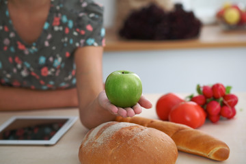 Close-up of female hand holding green apple in kitchen interiors. Many vegetables and other meal at glass table are ready for been cooked soon