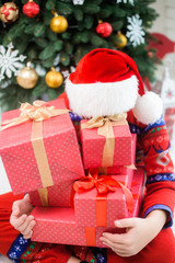 Funny little child in christmas red hat hiding behind pile of holiday presents. Green beautiful xmas tree in background. Vertical color photography.