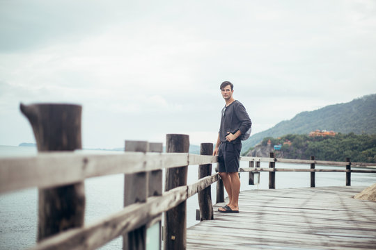 Landscape image of young man on wooden boardwalk by sea looking off into the distance