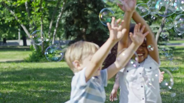 SLOWMO of laughing little African girl and three cute boys running and jumping on lawn in the park while catching soap bubbles in the air