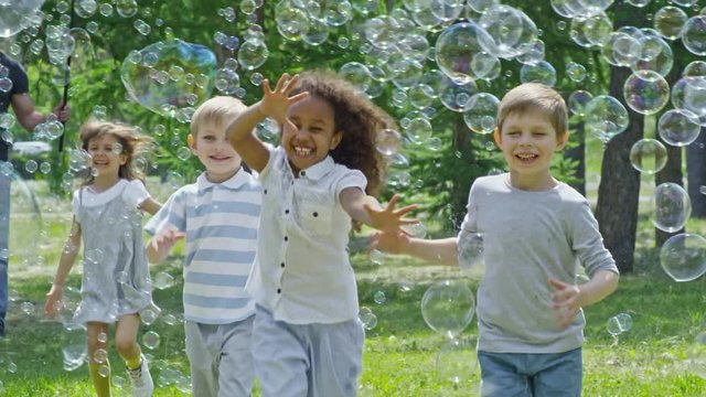 Tilt down of happy little kids running at camera, smiling and playing with lots of soap bubbles in park