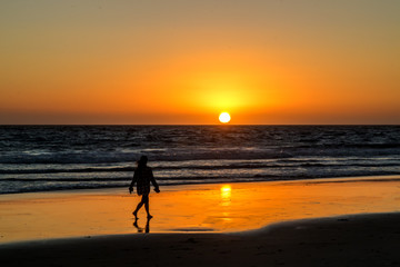 Fototapeta na wymiar Red, Yellow and Orange Sunset Over Wave covered Ocean with Woman Walking in Wet Sand Reflecting Sun