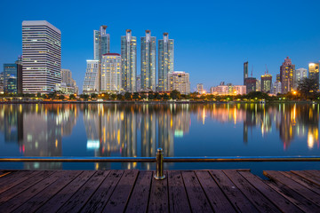 Naklejka premium Benjakiti Park in Bangkok, Thailand. In the night with calm water reflecting buildings, wooden walk way and blue sky background.